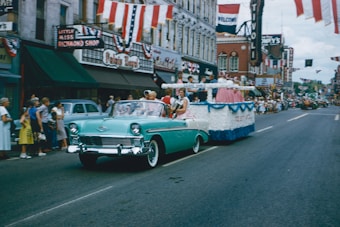 A vibrant parade scene on a busy street with a vintage turquoise convertible. People are riding in the car, followed by a festive float adorned with colorful decorations. The backdrop features shops with patriotic banners and many onlookers lining the street, adding a lively atmosphere to the event.