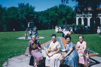 Several women sit on a circular metal structure in a park, smiling at the camera. In the background, children play on a jungle gym, while a group of people interact around picnic tables. A large building with arches is visible in the distance, surrounded by lush greenery under a clear blue sky.