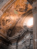 The image displays the intricately decorated ceiling of a historic building, possibly a church or cathedral. The ceiling features elaborate frescoes and reliefs depicting various scenes with multiple human figures. The artwork is rich in detail, with a warm color palette dominated by gold, earth tones, and subtle greens. An arched window allows natural light to illuminate part of the ceiling, enhancing the grandeur and depth of the artistic work.