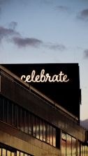 A modern building with a large sign on the roof displaying the word 'celebrate' in illuminated letters. The sky is partly cloudy, transitioning into evening with a soft glow reflecting on the building's glass windows.