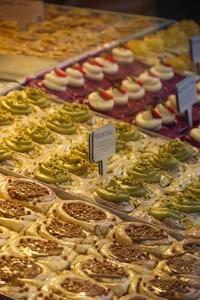 A display of assorted pastries, including pistachio rolls with green frosting and crushed nuts, cream-filled desserts topped with strawberries, and caramel-topped pastries. Arranged neatly in rows, each type has its own section. Small signage in the center indicates the type of pastry.