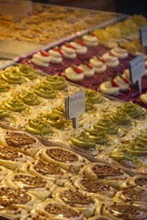 A display of assorted pastries, including pistachio rolls with green frosting and crushed nuts, cream-filled desserts topped with strawberries, and caramel-topped pastries. Arranged neatly in rows, each type has its own section. Small signage in the center indicates the type of pastry.