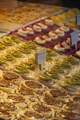 A display of assorted pastries, including pistachio rolls with green frosting and crushed nuts, cream-filled desserts topped with strawberries, and caramel-topped pastries. Arranged neatly in rows, each type has its own section. Small signage in the center indicates the type of pastry.