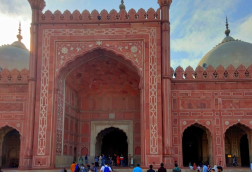 An ornate, historic red sandstone building with intricate floral and geometric designs. The structure has large archways and domes, showing Islamic architectural influence. People can be seen at the entrance, indicating it is a place of significance, possibly a mosque or historical landmark.