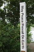 A tall, white pole inscribed with the phrase 'May Peace Prevail On Earth' in bold black letters stands amidst lush green foliage. The background is filled with dense greenery, suggesting a natural, serene setting.