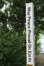 A tall, white pole inscribed with the phrase 'May Peace Prevail On Earth' in bold black letters stands amidst lush green foliage. The background is filled with dense greenery, suggesting a natural, serene setting.