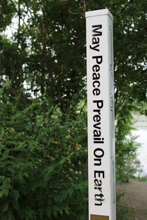 A tall, white pole inscribed with the phrase 'May Peace Prevail On Earth' in bold black letters stands amidst lush green foliage. The background is filled with dense greenery, suggesting a natural, serene setting.