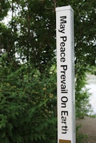 A tall, white pole inscribed with the phrase 'May Peace Prevail On Earth' in bold black letters stands amidst lush green foliage. The background is filled with dense greenery, suggesting a natural, serene setting.