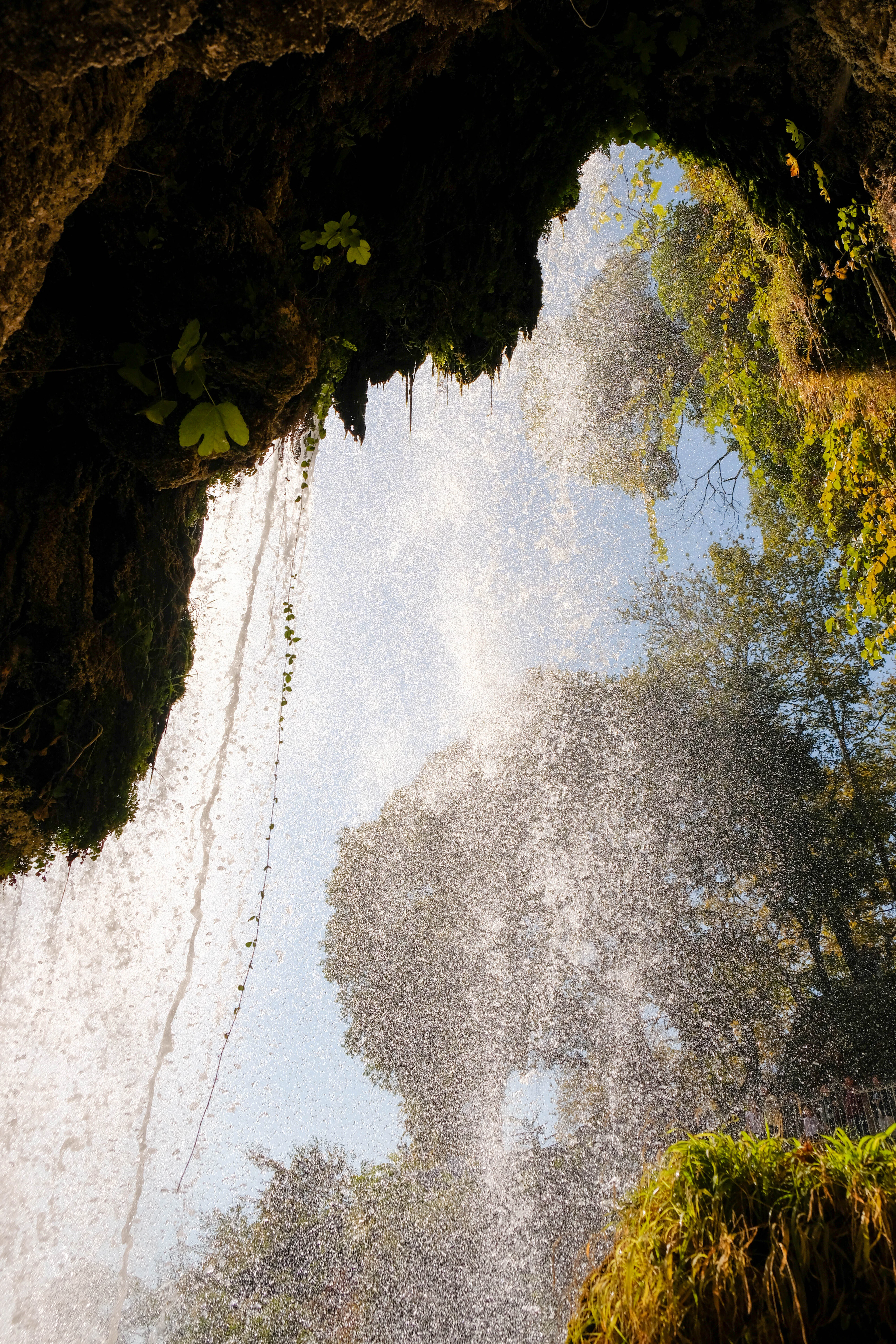 Looking out from the mossy cave mouth behind the Karanos waterfall at Edessa, sunlit spray falling in a bright curtain across the greenery beyond.