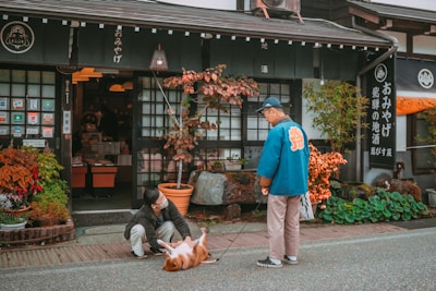 A man wearing a blue jacket and cap stands with a leash in front of a traditional Japanese store. A person sits on the ground next to a brown and white dog lying on its back. The shop has Japanese signage, and there are potted plants and colorful foliage around the entrance.