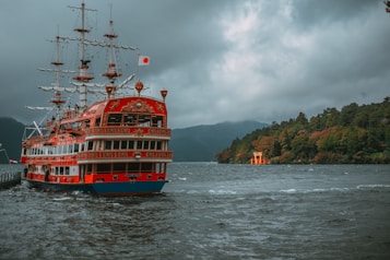 A large, ornate ship with multiple decks and masts is sailing on a river or lake. The ship is painted predominantly red with intricate designs and has a Japanese flag on top. In the background, tree-covered hills can be seen under an overcast sky. A traditional Japanese torii gate stands near the shore on the right side of the image.