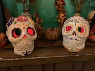 A pair of decorative sugar skulls are displayed on a dark wooden surface. Each skull features intricate designs with bright colors like purple, orange, green, and red. In the background, autumn leaves and small pumpkins add to the festive décor.