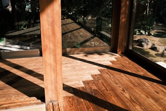 Warm sunlight casts geometric shadows on wooden flooring inside a modern structure, with large glass windows revealing a view of a garden outside. The natural textures of the wood are highlighted, creating a calm and serene environment.