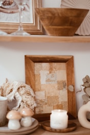 A cozy, rustic shelf is filled with decorative items, including a framed collage of vintage papers in warm tones, a dried floral wreath, a trio of ceramic mushroom-shaped ornaments, a lit candle on a wooden tray, and a large wooden bowl. The neutral color palette and natural textures create a warm and inviting atmosphere.
