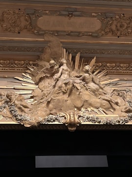 A detailed and ornate bas-relief sculpture depicting classical figures surrounded by decorative elements. The central figure seems to be seated or reclining, with other figures around. The sculpture includes rays or spiky elements extending outward and floral patterns that embellish the surrounding area. The phrase 'AD ALTA PER ARTES' is inscribed above the figures.