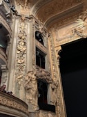 Detailed and ornate architectural interior with intricate golden designs. Sculptures and carvings decorate the walls, portraying human figures and floral patterns. Multiple small balconies are visible with people seated in them. The overall aesthetic is classical and opulent, with heavy use of gold and elaborate detailing.
