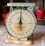 An antique, round kitchen scale with a cream-colored face and green accents is prominently displayed. The scale reads 'American Family Scale' and shows measurements in pounds and ounces. A red apple-shaped container sits in the background, along with stacked red bowls.