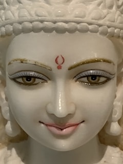 A close-up of a serene white marble statue with intricate facial features. The statue's eyes are delicately painted, and it has a small red symbol on the forehead. The lips are painted a soft pink, and the eyebrows are accentuated with golden highlights.