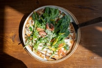 A freshly baked pizza topped with vibrant greens, cherry tomatoes, yellow bell peppers, and drizzled with balsamic glaze. The pizza is placed on a wooden table, with a light shining from the side creating a warm and inviting atmosphere.
