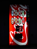 A well-lit vending machine displays several beverage options, including bottled drinks such as water, soda, and juices. The machine's bright red and white design features the Coca-Cola logo prominently.