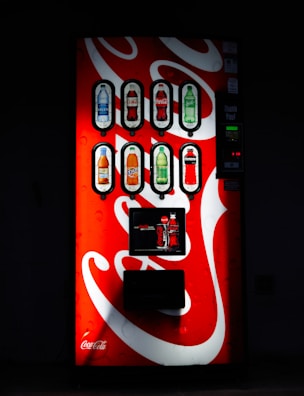A well-lit vending machine displays several beverage options, including bottled drinks such as water, soda, and juices. The machine's bright red and white design features the Coca-Cola logo prominently.