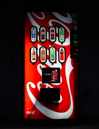 A well-lit vending machine displays several beverage options, including bottled drinks such as water, soda, and juices. The machine's bright red and white design features the Coca-Cola logo prominently.