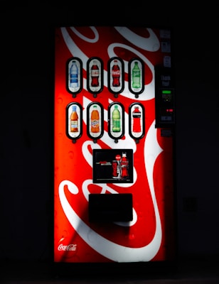 A well-lit vending machine displays several beverage options, including bottled drinks such as water, soda, and juices. The machine's bright red and white design features the Coca-Cola logo prominently.