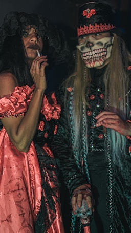 Two individuals dressed in elaborate costumes, one wearing a long black dress with red lace and a large black hat, while holding a cigarette. The other person is wearing a detailed skeleton mask with a long-haired wig and a decorated top hat. Both are dressed in ornate, theatrical black and red outfits, conveying a gothic or Halloween theme.