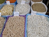 Various types of legumes and grains are displayed in bags at a market. Each bag is marked with a handwritten price sign. The bags include beans, lentils, and chickpeas, among others.