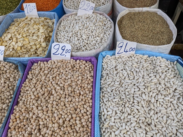 Various types of legumes and grains are displayed in bags at a market. Each bag is marked with a handwritten price sign. The bags include beans, lentils, and chickpeas, among others.