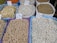 Various types of legumes and grains are displayed in bags at a market. Each bag is marked with a handwritten price sign. The bags include beans, lentils, and chickpeas, among others.