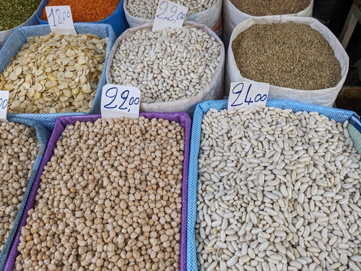 Various types of legumes and grains are displayed in bags at a market. Each bag is marked with a handwritten price sign. The bags include beans, lentils, and chickpeas, among others.