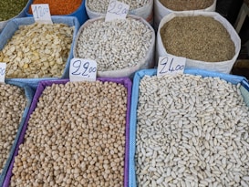Various types of legumes and grains are displayed in bags at a market. Each bag is marked with a handwritten price sign. The bags include beans, lentils, and chickpeas, among others.