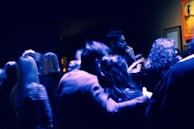 A group of people gathered in a dimly lit space, likely an art gallery or event. The lighting casts a blue hue over the scene, with some framed artwork visible on the walls. The atmosphere appears casual and social as individuals are engaged in conversation.