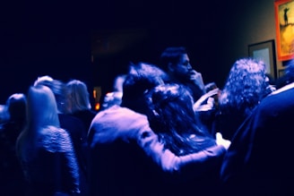 A group of people gathered in a dimly lit space, likely an art gallery or event. The lighting casts a blue hue over the scene, with some framed artwork visible on the walls. The atmosphere appears casual and social as individuals are engaged in conversation.