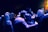 A group of people gathered in a dimly lit space, likely an art gallery or event. The lighting casts a blue hue over the scene, with some framed artwork visible on the walls. The atmosphere appears casual and social as individuals are engaged in conversation.
