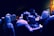 A group of people gathered in a dimly lit space, likely an art gallery or event. The lighting casts a blue hue over the scene, with some framed artwork visible on the walls. The atmosphere appears casual and social as individuals are engaged in conversation.