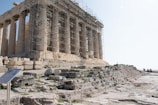 An ancient temple partially undergoing restoration, with scaffolding covering part of the structure. The temple is made of stone, featuring classical columns and surrounded by scattered ruins and stone steps. A few people are visible in the distance, adding a sense of scale.