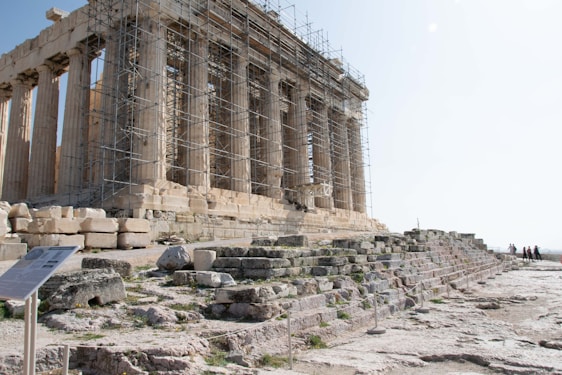 An ancient temple partially undergoing restoration, with scaffolding covering part of the structure. The temple is made of stone, featuring classical columns and surrounded by scattered ruins and stone steps. A few people are visible in the distance, adding a sense of scale.