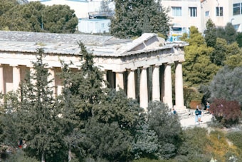 An ancient Greek temple with tall Doric columns is surrounded by lush green trees. The structure appears weathered, with a stone roof and a pediment at one end. People are walking around the temple, indicating it might be a historical or tourist site. Modern buildings are visible in the background.