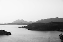 A serene monochrome landscape showcasing a vast body of water in the foreground with a rugged coastline extending into the distance. Rolling hills and mountains provide a striking backdrop, creating a harmonious contrast between the water and the land. The scene is peaceful and uninhabited, emphasizing the natural beauty of the terrain.