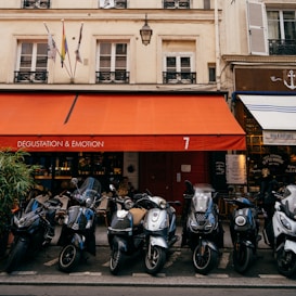 A row of scooters parked in front of a quaint French café with an orange awning and 'Dégustation & Émotion' written on it. Several flags are displayed above the café, and there are people sitting inside. The building has a classic European architectural style with shuttered windows.