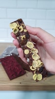 A hand is holding a chocolate bar topped with what appears to be dried fruit, such as banana slices and raisins. The bar has a smooth surface with some textured irregularities due to the toppings. In the background on a wooden surface lies another chocolate bar, partially wrapped, featuring a red topping.