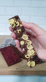 A hand is holding a chocolate bar topped with what appears to be dried fruit, such as banana slices and raisins. The bar has a smooth surface with some textured irregularities due to the toppings. In the background on a wooden surface lies another chocolate bar, partially wrapped, featuring a red topping.