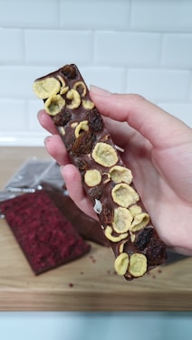 A hand is holding a chocolate bar topped with what appears to be dried fruit, such as banana slices and raisins. The bar has a smooth surface with some textured irregularities due to the toppings. In the background on a wooden surface lies another chocolate bar, partially wrapped, featuring a red topping.