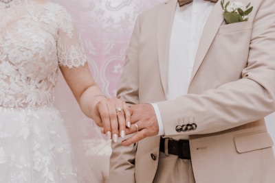 A couple standing close together, showcasing their hands with wedding rings. The woman is wearing a white lace wedding dress, and the man is dressed in a beige suit with a white shirt and bow tie. A boutonnière with white flowers is pinned on the man's lapel.