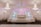 An elegantly decorated event hall featuring grand chandeliers and white floral arrangements. The symmetrical design includes an archway with soft pink lighting and a stage area adorned with decorative columns and flowers. Tables with white chairs are arranged along the sides.