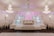 An elegantly decorated event hall featuring grand chandeliers and white floral arrangements. The symmetrical design includes an archway with soft pink lighting and a stage area adorned with decorative columns and flowers. Tables with white chairs are arranged along the sides.