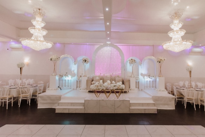 An elegantly decorated event hall featuring grand chandeliers and white floral arrangements. The symmetrical design includes an archway with soft pink lighting and a stage area adorned with decorative columns and flowers. Tables with white chairs are arranged along the sides.