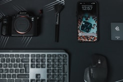 a keyboard, mouse, camera, and cell phone on a desk